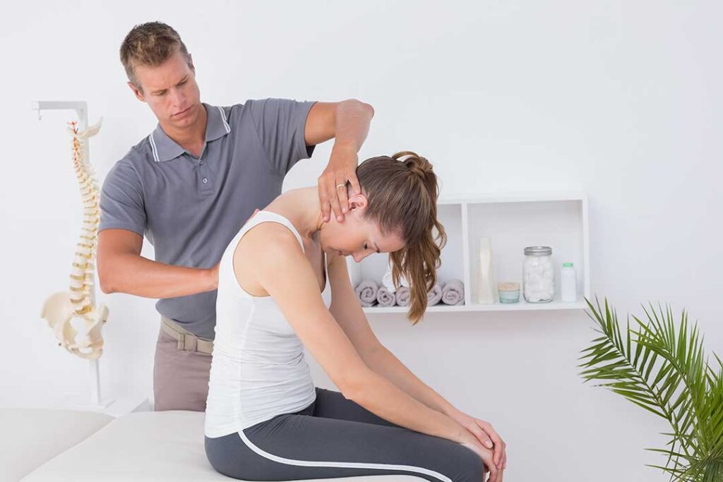 A pregnant woman stands in a living room, stretching gently with one hand on her lower back.